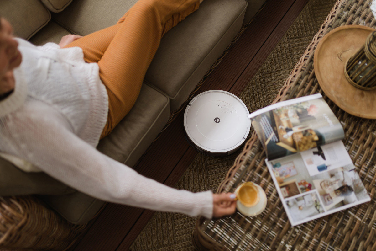 Starsza kobieta odpoczywa z kawą i gazetą na kanapie, przed kanapą sprząta robot iRobot Roomba