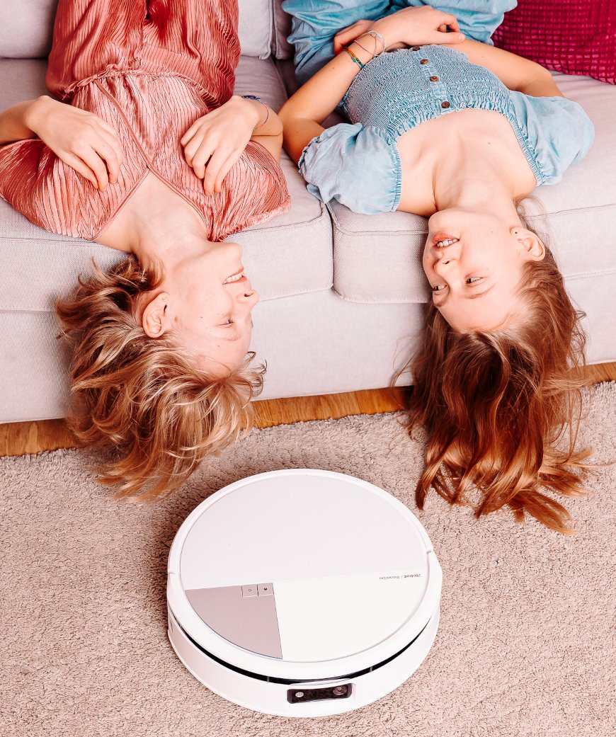 Dwie dziewczynki leżą na kanapie w salonie zwisając głową w dół, przed kanapą Roomba Max 705 Combo odkurza dywan