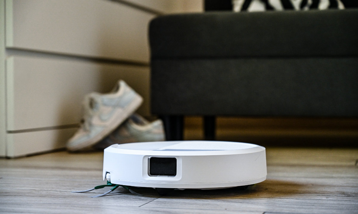 Robot Roomba sprząta salon, na drugim planie sofa i trampki