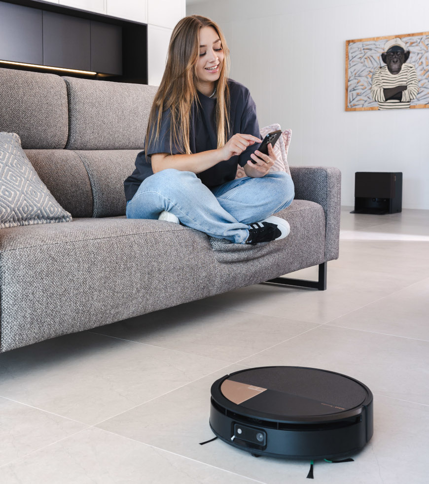 Kobieta siedzi na szarej sofie w salonie i trzyma telefon, Roomba 705 Combo sprząta kafle, na drugim planie stacja AutoWash