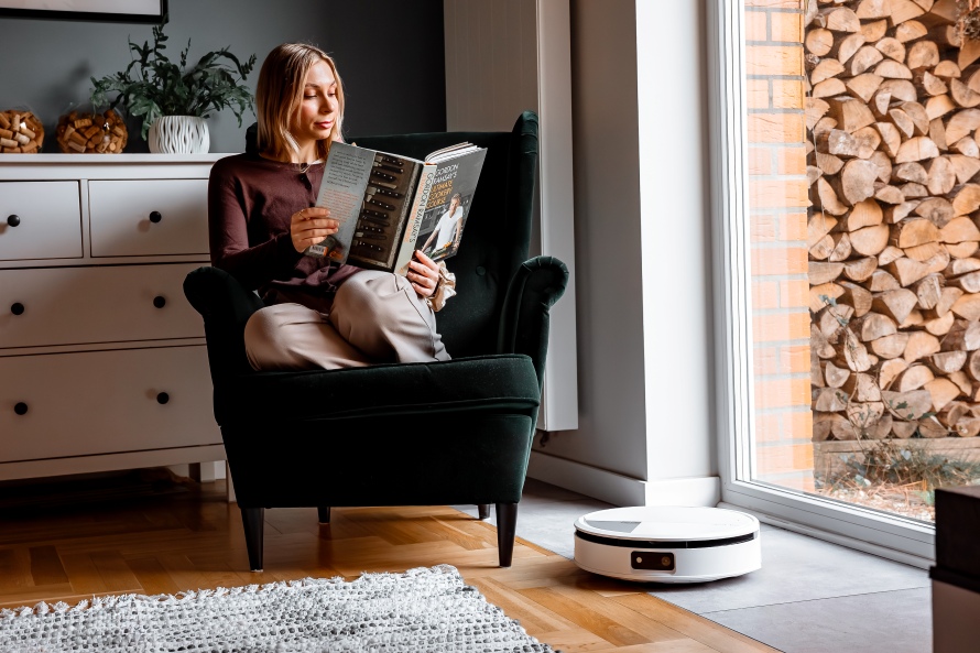 Kobieta czyta książkę w fotelu przy dużym oknie balkonowym, obok niej robot Roomba Max 705 Combo sprząta podłogę