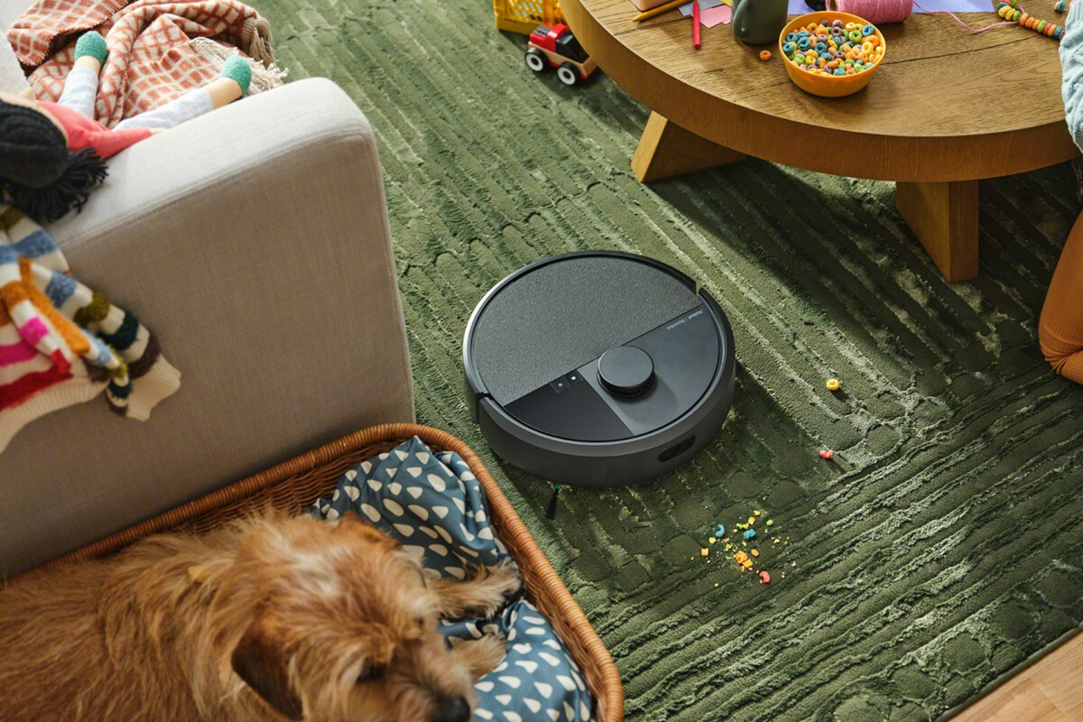 Roomba 405 podczas odkurzania dywanu w salonie. Obok pies leżący w legowisku