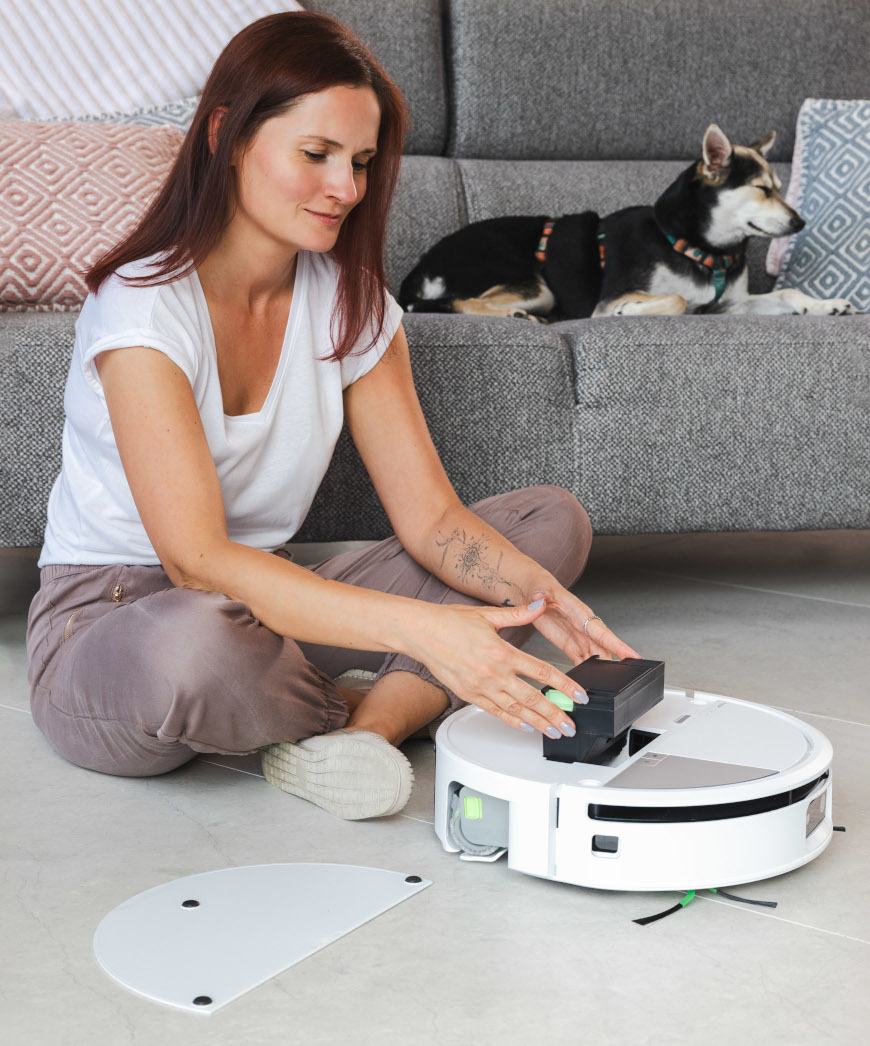 Kobieta wkłada pojemnik na brud do robota Roomba Max 705 Combo, na drugim planie pies leżący na szarej sofie