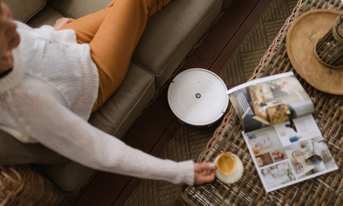 Zadowolona kobieta leży na kanapie i sięga po filiżankę kawy, podczas gdy na podłodze Roomba sprząta.