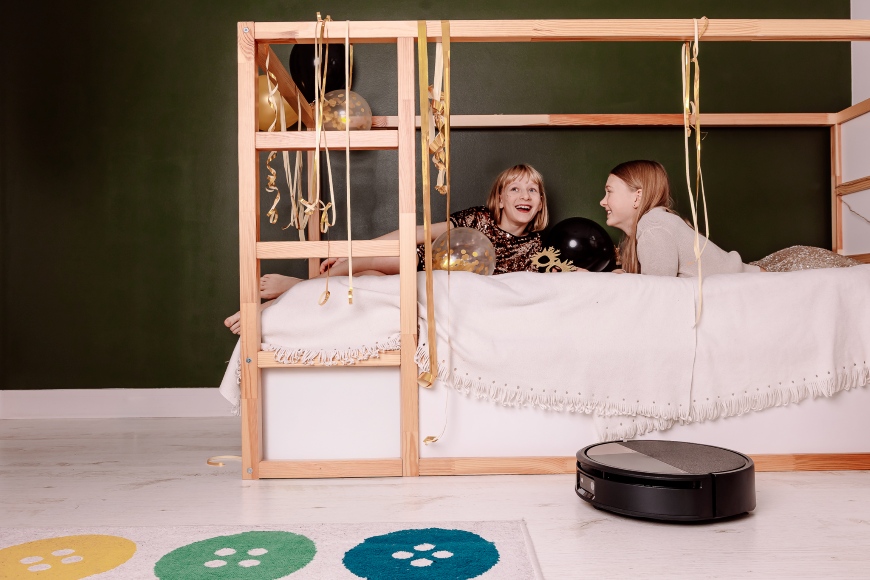 Dwie dziewczynki świętujące sylwestra leżą na łóżku, z którego zwisają ozdoby, na pierwszym planie sprząta Roomba Max 705 Combo