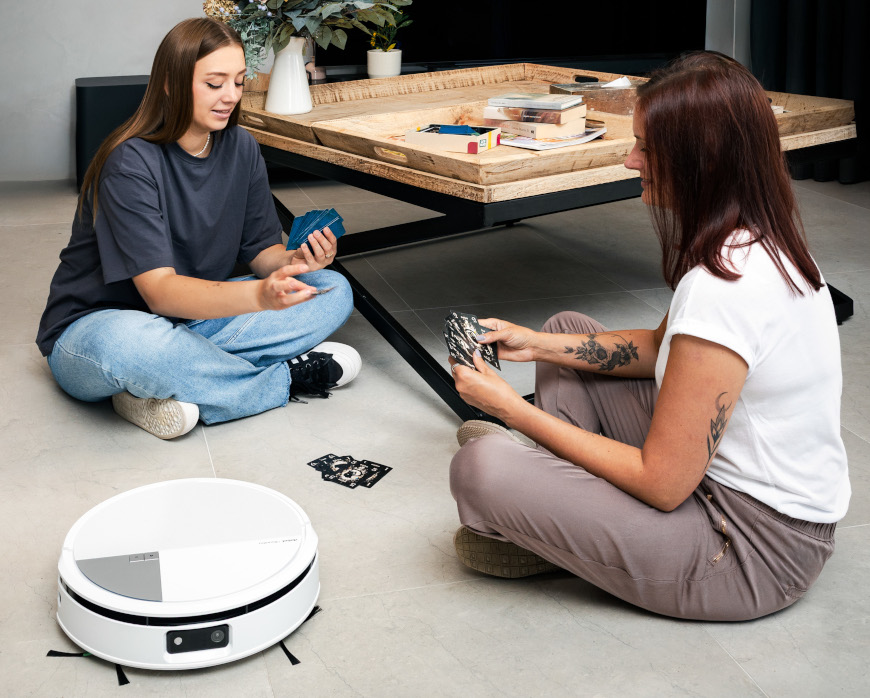 Dwie kobiety grają w karty na podłodze w salonie przy stoliku kawowym, obok nich sprząta robot Roomba Max 705 Combo