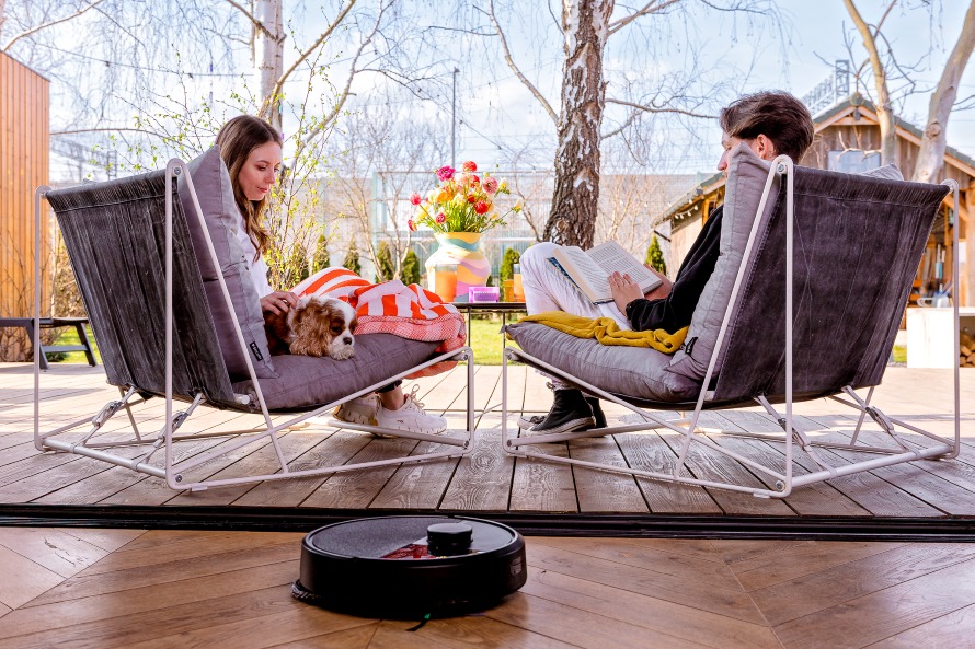 Para z psem siedzi na tarasie z widokiem na ogród. Na pierwszym planie Roomba Plus 505 Combo odkurza i mopuje twardą podłogę