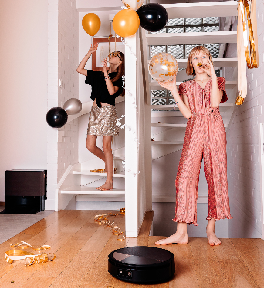 Dziewczynki bawią się balonami przy schodach, wokół konfetti, na pierwszym planie sprząta Roomba Max 705 Combo