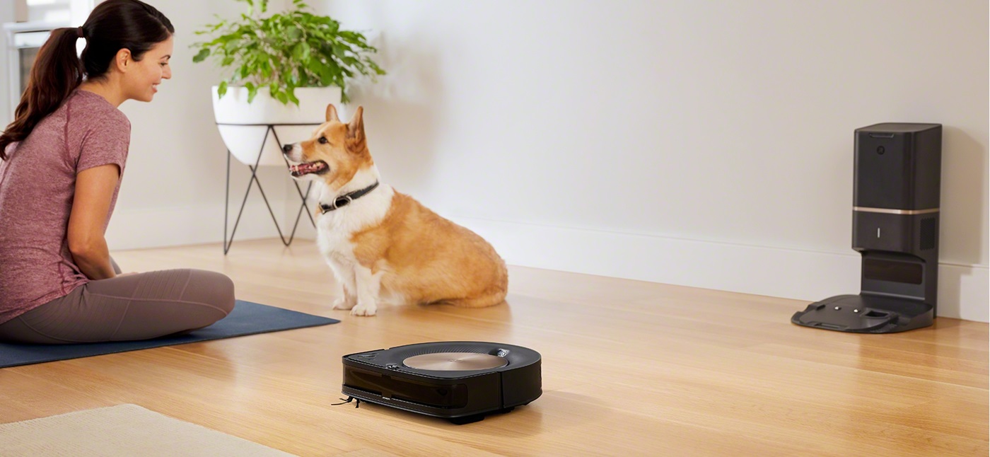 iRobot Roomba s9 wyjeżdża ze stacjo dokującej, w tle pies i kobieta