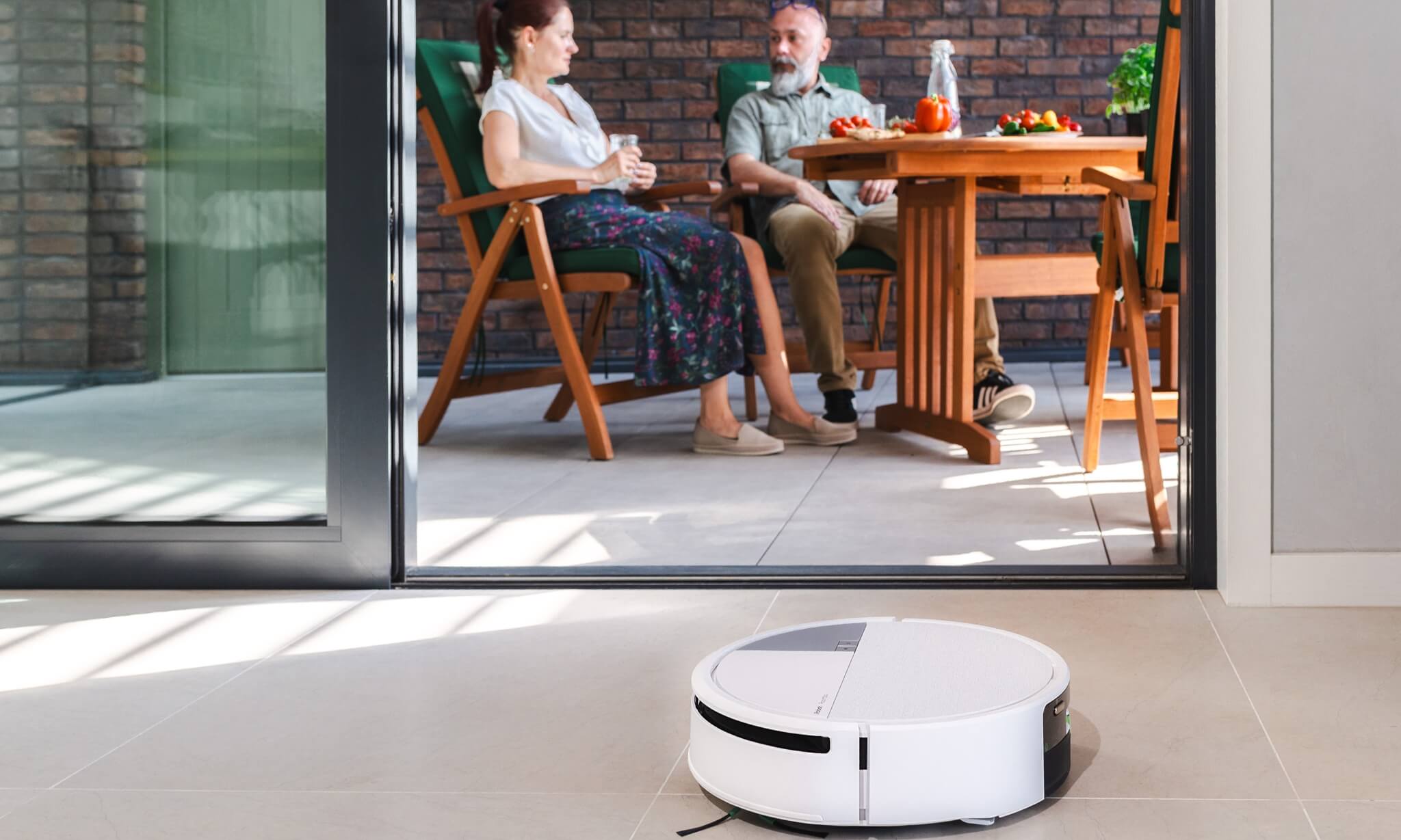 Małżeństwo je lunch na tarasie, na pierwszym planie robot Roomba Max 705 Combo sprząta marmurową podłogę