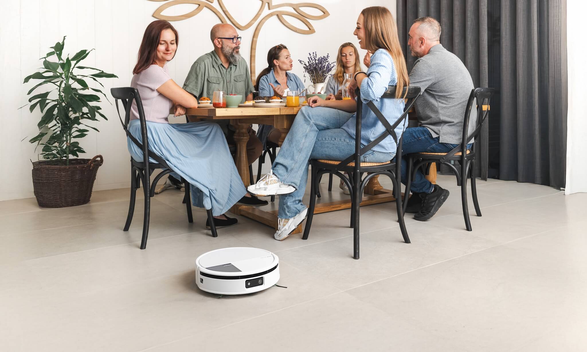 Rodzina je śniadanie przy dużym stole w jadalni, kobieta patrzy na sprzątającego robota Roomba Max 705 Combo