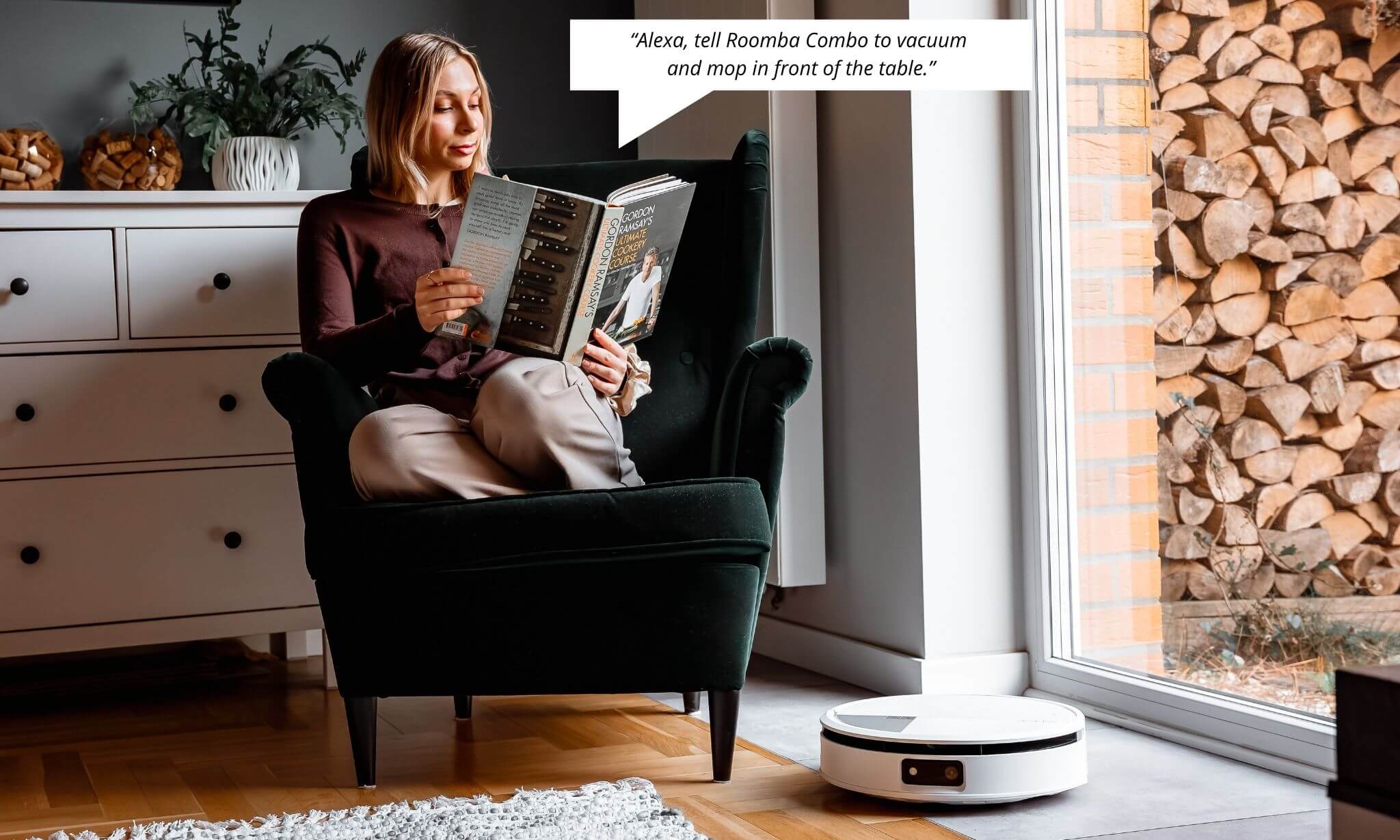 Kobieta czyta książkę siedząc w zielonym fotelu obok komody i okna balkonowego, na pierwszym planie sprząta iRobot Roomba Combo