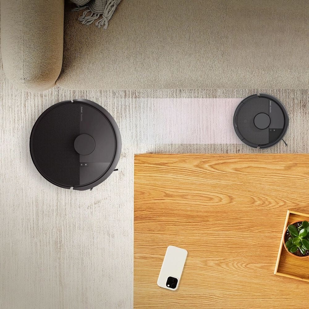 Robot Roomba Mini przejeżdża wąską ścieżką między kanapą a stolikiem kawowym, obok Roomba 105 Combo