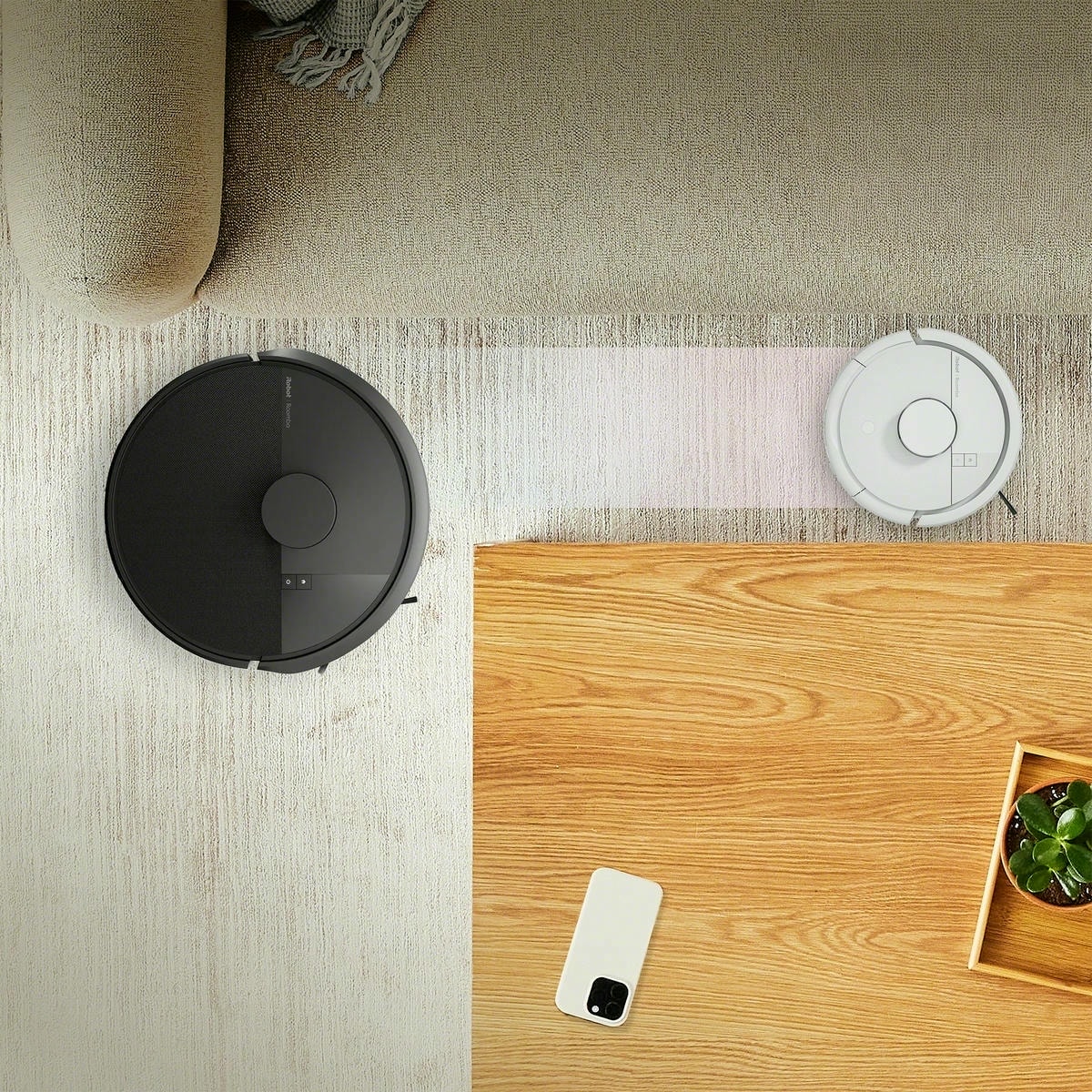 Robot Roomba Mini przejeżdża wąską ścieżką między kanapą a stolikiem kawowym, obok Roomba 105 Combo