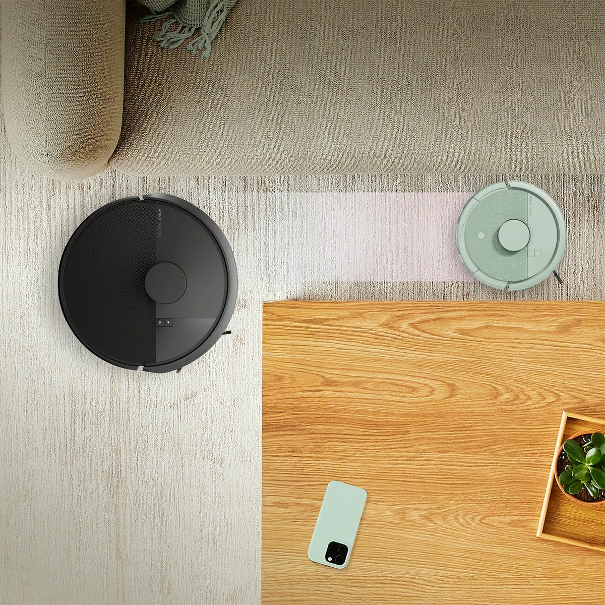Robot Roomba Mini przejeżdża wąską ścieżką między kanapą a stolikiem kawowym, obok Roomba 105 Combo