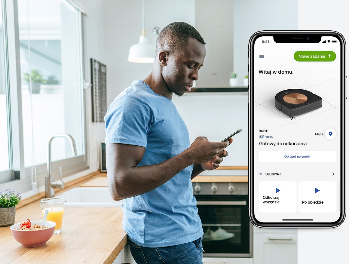 Mężczyzna stoi w kuchni. Trzyma w ręku smartfona. Przegląda aplikację iRobot Home.